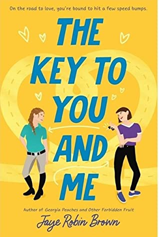 Book cover of The Key to You and Me. Two characters stand in front of a yellow background with a road pattern shaped into a heart. The character on the left has a red braid, a green t shirt, khaki pants, and black high riding boots. The character on the right has a brown bob, a purple shirt, black skinny jeans, and purple sneakers. She is holding car keys and an arrow points from them to the girl on the left. 